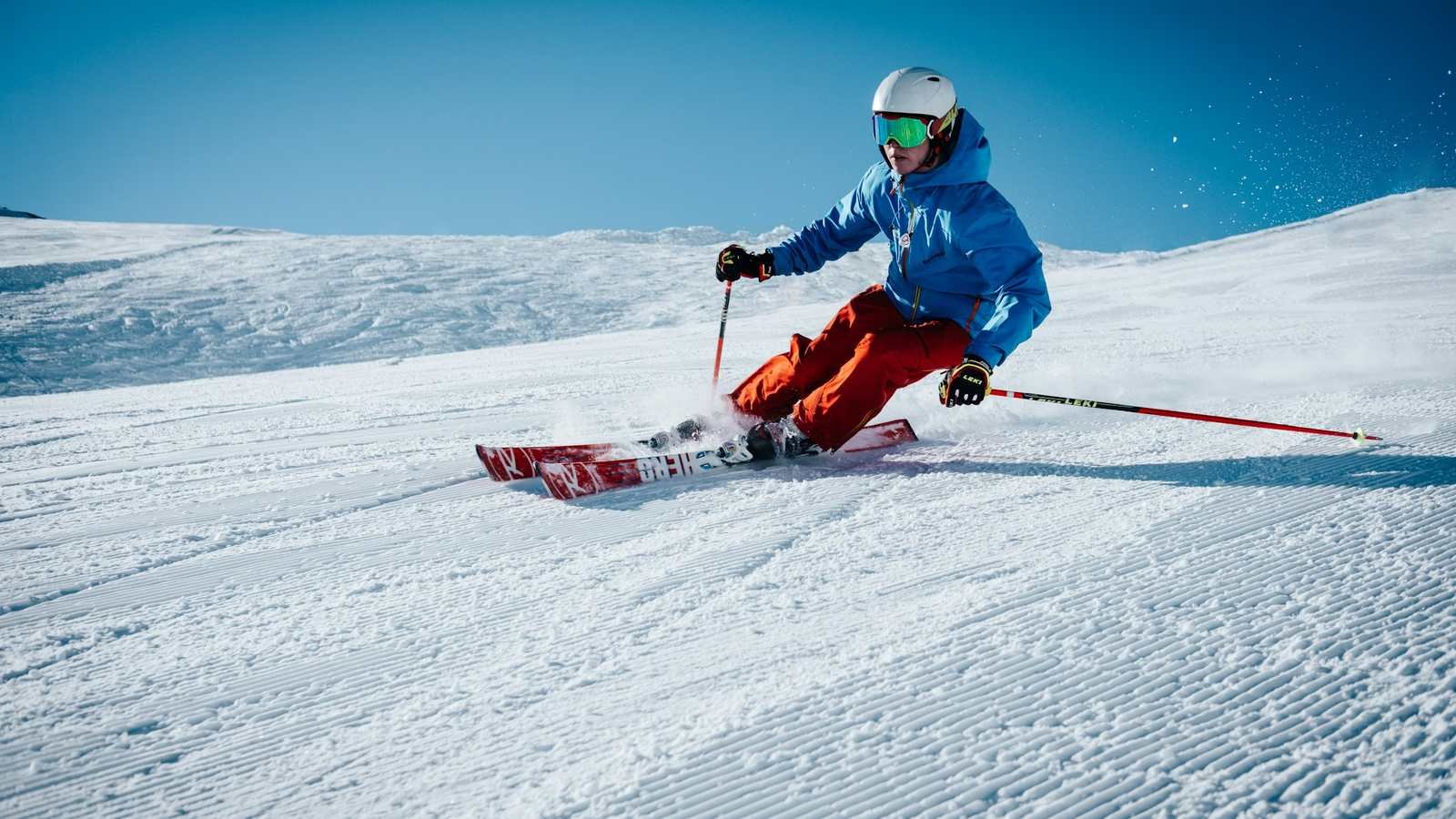 A skier in a blue jacket carving down a groomed piste on a clear day